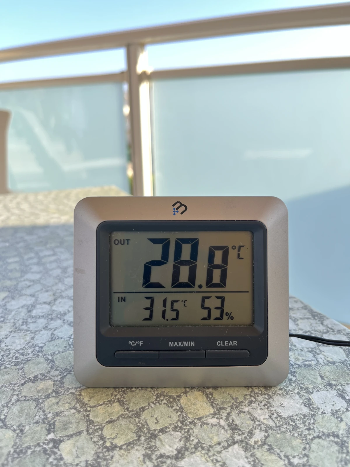 Sisä- ja ulkolämpötilaa näyttävä mittari pöydällä, taustalla kaide. Lämpötila 28.8 astetta.